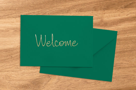Welcome greeting handwritten on a green envelope and card lying on a wood table viewed from above