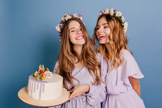Beautiful brunette girl in purple dress holding birthday cake. Indoor portrait of two ladies with flower in hair enjoying party together.