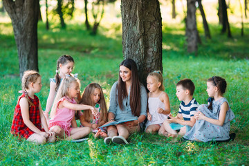 Fototapeta premium Children and education, young woman at work as educator reading book to boys and girls in park.