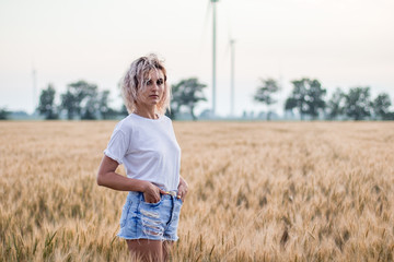 young tanned curly haired woman in white t-short and denim shorts is happy to be on the field of ripened wheat, wind turbine on the background