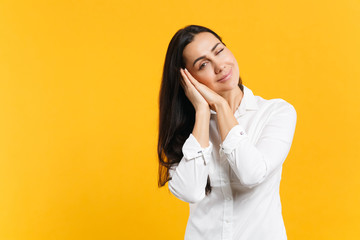 Portrait of funny blinking young woman in white casual shirt sleep with folded hands under cheek isolated on yellow orange wall background in studio. People lifestyle concept. Mock up copy space.