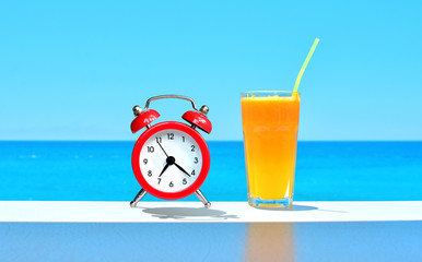 Vacation background. It's time to travel concept. It's time to rest. Orange fresh juice and red alarm clock standing on the table on a sea water backdrop.