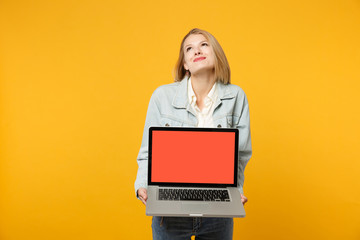 Pensive young woman in denim casual clothes looking up, holding laptop pc computer with blank empty screen isolated on yellow orange background in studio. People lifestyle concept. Mock up copy space.