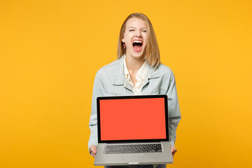 Portrait of crazy screaming young woman in denim casual clothes holding laptop pc computer with blank empty screen isolated on yellow orange background. People lifestyle concept. Mock up copy space.