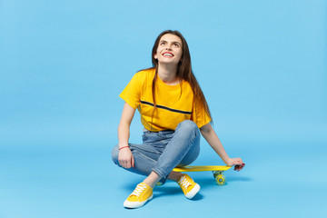 Portrait of cheerful cute young woman in vivid casual clothes looking up and sitting with yellow skateboard isolated on blue wall background in studio. People lifestyle concept. Mock up copy space.