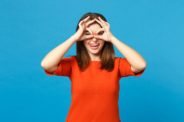 Beautiful woman wearing red orange dress holding hands near eyes, imitating glasses or binoculars isolated over blue background, studio portrait. People lifestyle fashion concept. Mock up copy space.