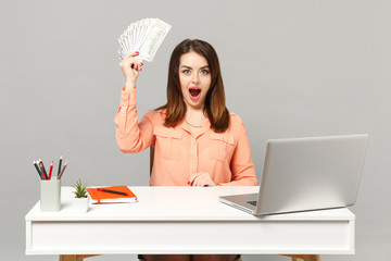 Young surprised woman holding fan of cash money in dollar banknotes, sit and work at desk with pc laptop isolated on gray background. Achievement business career lifestyle concept. Mock up copy space.