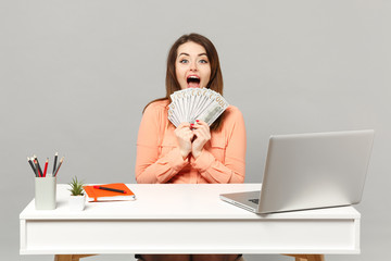 Young excited woman holding fan of cash money in dollar banknotes, sit and work at desk with pc laptop isolated on gray background. Achievement business career lifestyle concept. Mock up copy space.