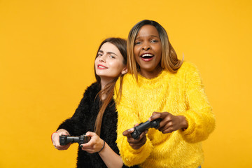 Two fun young women friends european and african american in black yellow clothes hold joystick isolated on bright orange wall background, studio portrait. People lifestyle concept. Mock up copy space © ViDi Studio