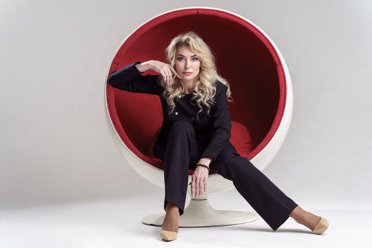 Image of blonde woman with long curly hair in black suit and beige shoes looking in camera while sitting in round chair isolated on white background