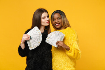 Two fun young women friends european and african in black yellow clothes holding fan of cash money...