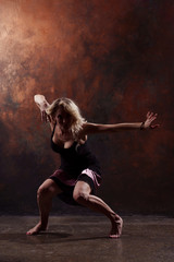 Photo of young dancing blonde with hands to sides in black dress on brown background
