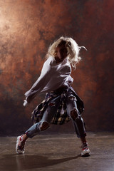 Photo of long-haired blonde dancer woman with developing hair in torn jeans and sneakers on dark brown background