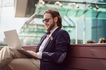 Photo on side of serious businessman with laptop sitting on wooden bench in city center on summer day