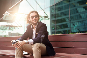 Photo of brunet with beard looking to side with glass in hand sitting on wooden bench against background of glass building