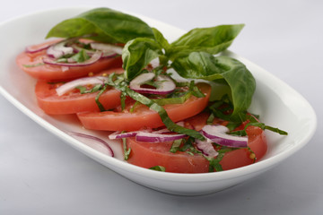 fresh tomato sliced ​​with ringlets with basil leaves and onions. On an oval white plate on a white background.