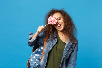 Young african american girl teen student in denim clothes, backpack hold candy isolated on blue wall background studio portrait. Education in high school university college concept. Mock up copy space