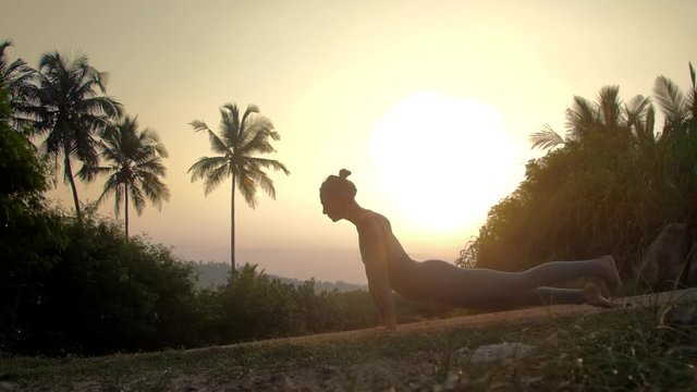 young lady silhouette gets into downward facing dog asana on track against morning back sunshine side view slow motion. Concept fitness yoga wellness lifestyle