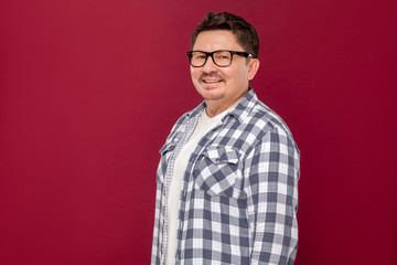 Portrait of happy successful handsome middle aged business man in casual checkered shirt and eyeglasses standing and looking at camera with toothy smile. studio shot, isolated on dark red background.