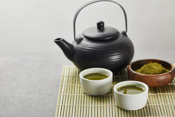 selective focus of green matcha powder in wooden bowl near black teapot and cups with tea on bamboo table mat