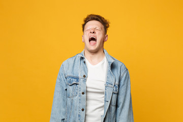 Portrait of crazy screaming young man in denim casual clothes keeping eyes closed isolated on yellow orange wall background in studio. People sincere emotions, lifestyle concept. Mock up copy space.