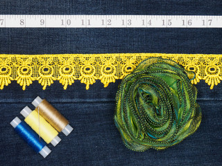 Yellow lace, threads with needle, tape measure and green bow on a jeans background. Sewing still...