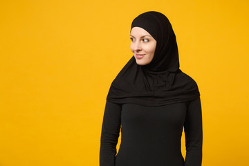 Smiling beautiful young arabian muslim woman in hijab black clothes looking aside camera isolated on yellow wall background, studio portrait. People religious lifestyle concept. Mock up copy space.