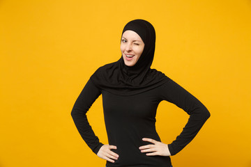 Young arabian muslim woman in hijab black clothes standing with arms akimbo on waist isolated on yellow wall background, studio portrait. People religious Islam lifestyle concept. Mock up copy space.