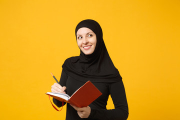 Young arabian muslim woman in hijab black clothes hold notebook, writing, prepare to exam isolated on yellow wall background, studio portrait. People religious lifestyle concept. Mock up copy space.