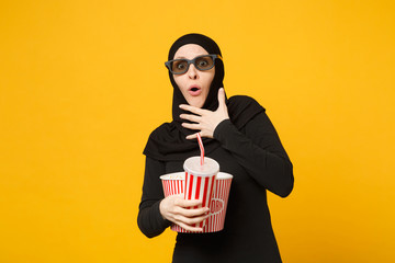 Arabian muslim woman in hijab black clothes 3d imax glasses watch movie film hold popcorn, cup of soda isolated on yellow wall background studio portrait. People lifestyle concept. Mock up copy space.
