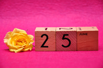 25 June on wooden blocks with a yellow rose on a pink background