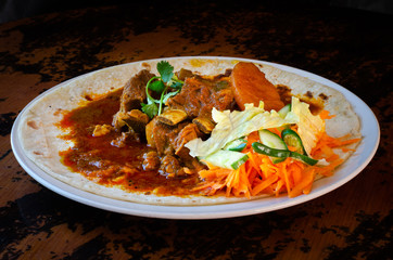 A mutton curry roti with sambals - an iconic Durban meal consisting of a roti topped with mutton curry and gravy. The sambals are grated carrot with chopped chilli.