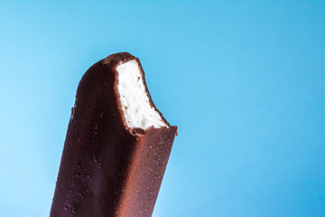 Bitten ice cream lolly in chocolate glaze close-up with condensate and icing frost on a summer blue sky background. Ice cream texture.