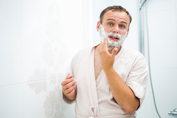 Young man applies shaving foam on face; looks in frame; prepares skin for shaving