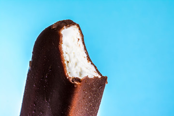 Bitten ice cream lolly in chocolate glaze close-up with condensate and icing frost on a summer blue sky background. Ice cream texture.