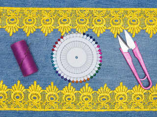 Yellow lace, thread with a needle, pins, scissors on a jeans background. Sewing still life