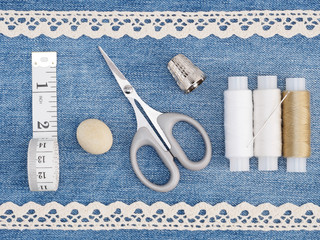 White lace, threads with needle, thimble, tape measure and scissors on jeans background. Stitching...