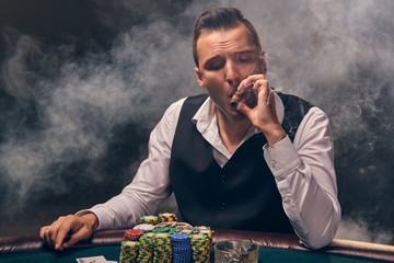 Wealthy man is smoking a cigar and playing poker with an excitement at a casino on black background.