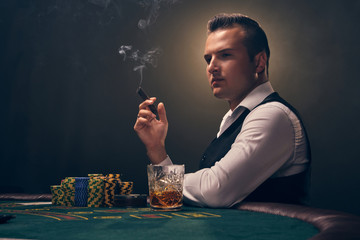 Wealthy man is smoking a cigar and playing poker with an excitement at a casino on black background.