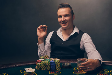 Wealthy man is smoking a cigar and playing poker with an excitement at a casino on black background.