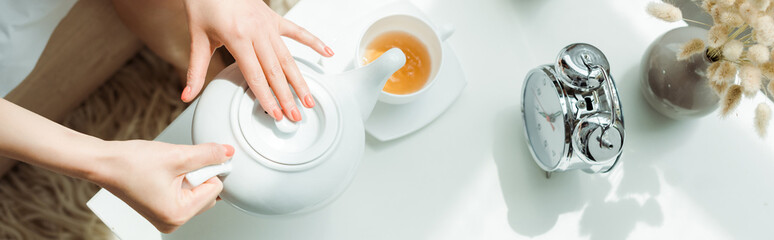 panoramic shot of woman holding teapot near cup and vintage alarm clock