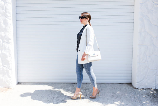 Pretty young fashion sensual woman walking by white wall background dressed in hipster style jeans and white jacket.