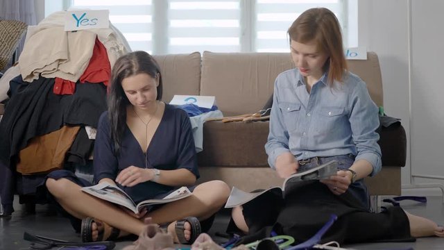 two female friends are sitting together on floor of living room and viewing magazines, flipping pages and chatting