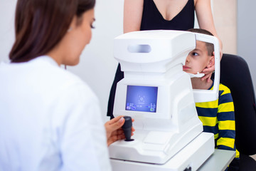 Female oculist using machine for checking eye sight in clinic. Little boy looking at equipment and...