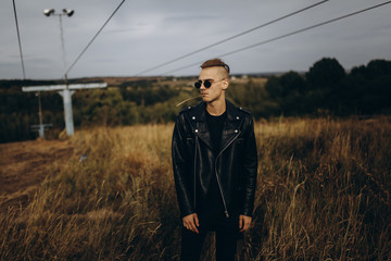 Young sexy man in leather jacket and sunglasses standing outdoor. Dark key photo