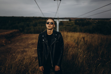 Young sexy man in leather jacket and sunglasses standing outdoor. Dark key photo