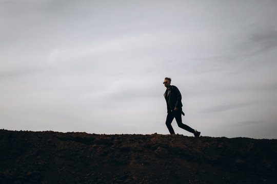 Young Sexy Running Man In Leather Jacket And Sunglasses Standing Outdoor. Silhouette Against Grey Sky