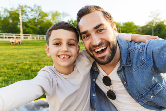 Young man having fun with his little brother or son outoors in park beautiful green grass take a selfie by camera.