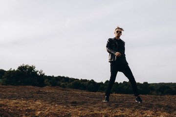 Young sexy man in leather jacket and sunglasses standing outdoor. Silhouette against grey sky