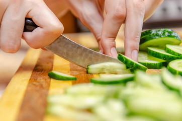 Female hand cuts cucumbers with a knife on the bars on a wooden plank close up